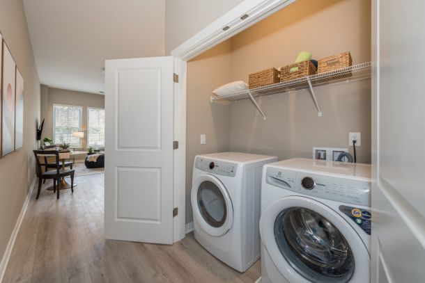 Attain on 5th apartment homes with Modern laundry closet with washer, dryer, wire shelf, and open door leading to a bright living area.