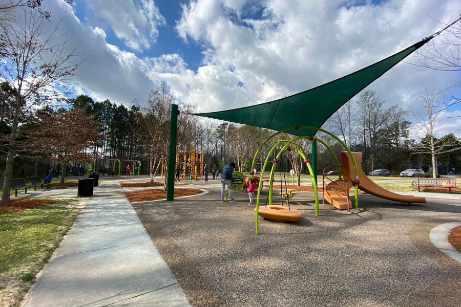 Acclaim at Cary Pointe apartment homes with Children and adults play at a colorful playground under a green shade on a partly cloudy day.