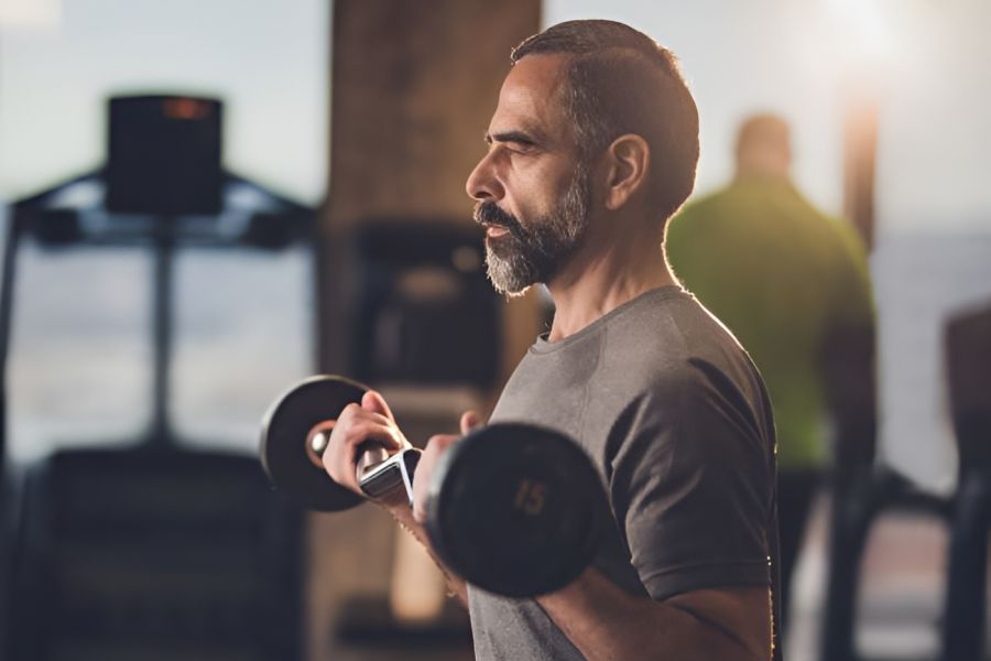 Acclaim at Cary Pointe apartment homes with Middle-aged man in a gym lifting dumbbells, wearing athletic clothes, with sunlight in the background.