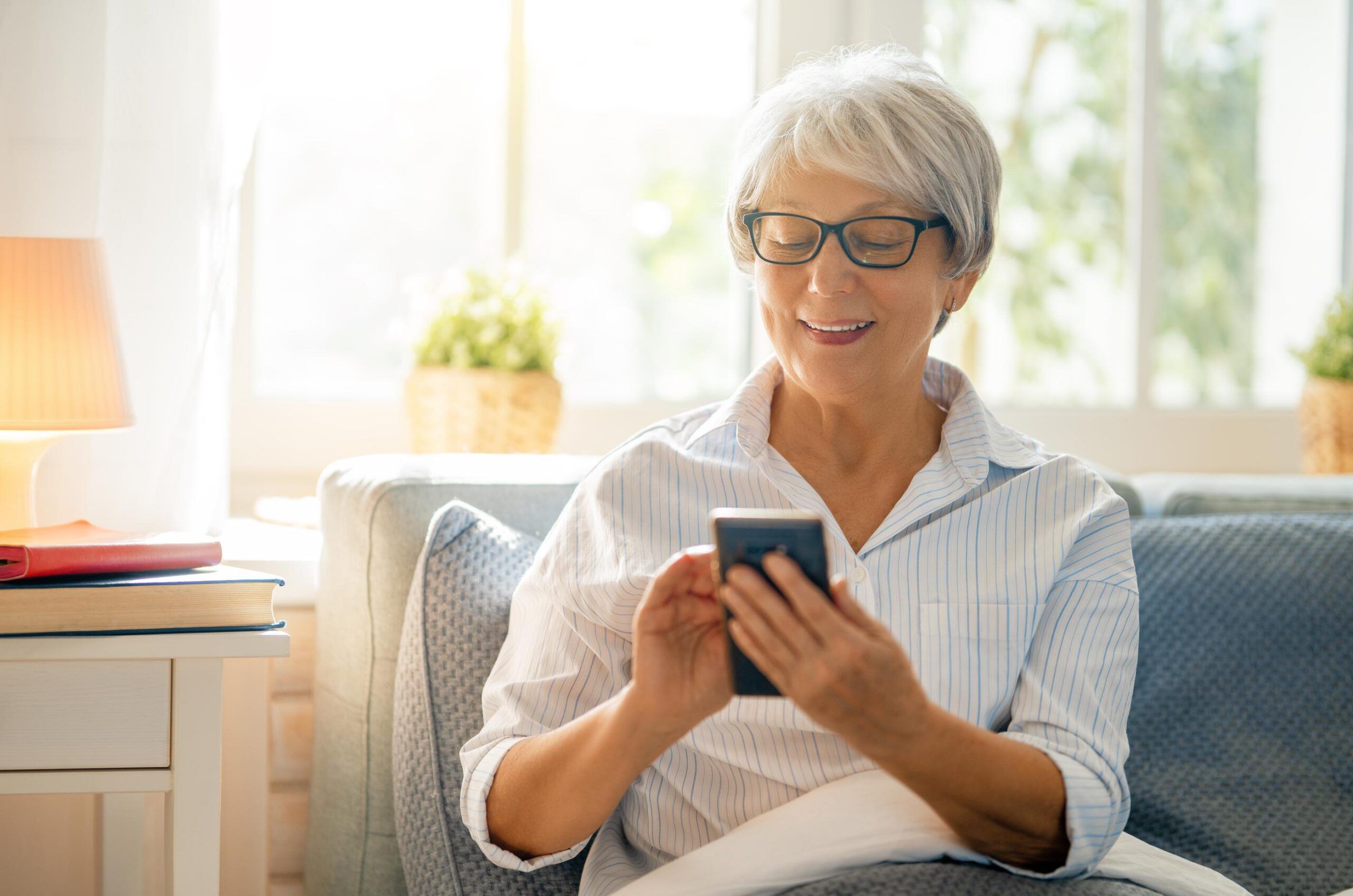 Attain on 5th apartment homes with Smiling older woman with glasses sits on a couch using a smartphone in a bright, cozy living room.