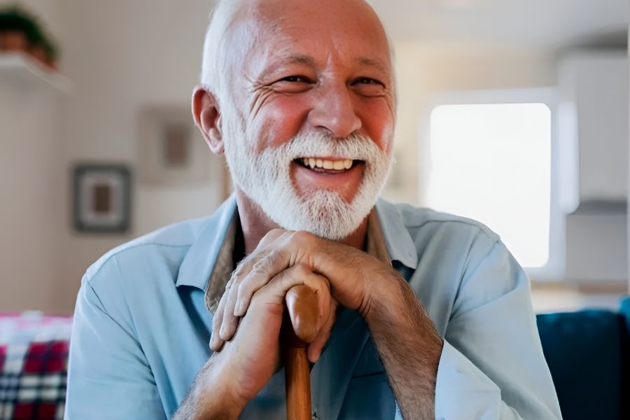 Acclaim at Cary Pointe apartment homes with Smiling older man with white hair and beard sits indoors, leaning on a wooden cane, wearing a light blue shirt.