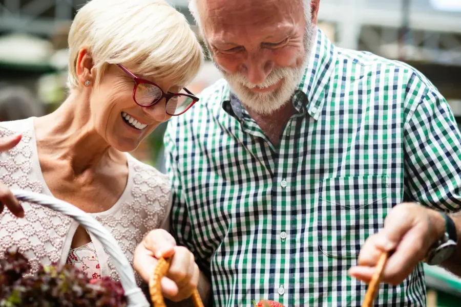 Cedar Grove Smiling elderly couple shopping at an outdoor market, holding baskets of fresh produce.